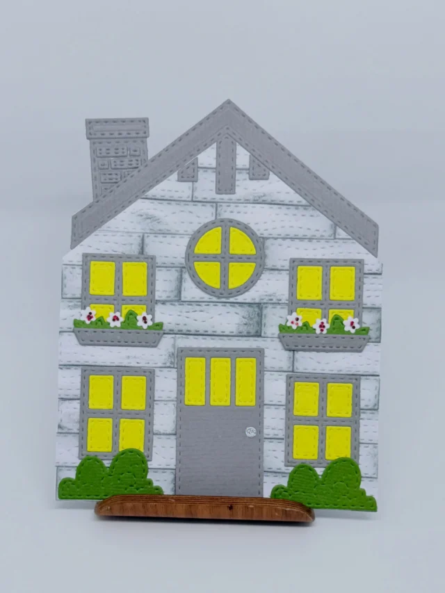 Karte mit grauem Papierhaus, gelb leuchtenden Fenstern, Blumenkästen und Büschen