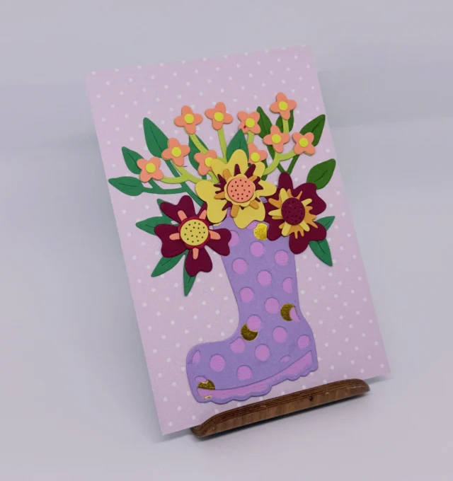Rosa Karte mit einem lila gepunkteten Gummistiefel, aus dem Papierblumen wachsen