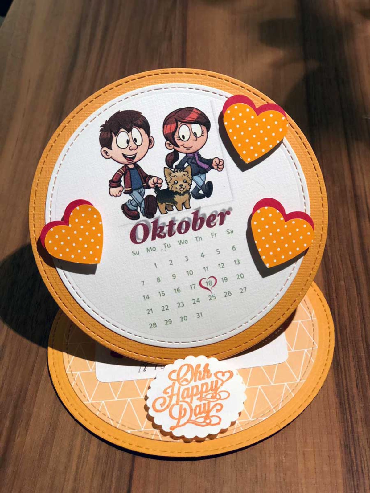 Runde Glückwunschkarte in Orange mit Kalenderblatt, Herzen und Comic-Figuren